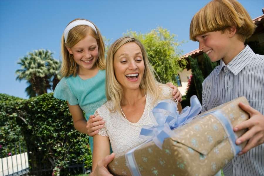 Mère blonde surprise et ravie recevant une grande boîte cadeau fête des mères kraft à ruban bleu de ses deux enfants souriants en plein air.