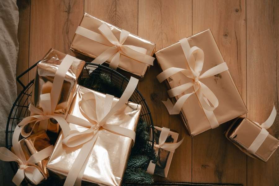 Une collection de cadeaux originaux emballés dans du papier doré brillant et noués avec des rubans crème, posés sur un plancher en bois.
